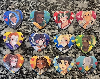 Puede incluir: Una colección de botones en forma de corazón con ilustraciones coloridas de varios personajes. Cada botón tiene un diseño único con diferentes combinaciones de colores y retratos de personajes. Los botones están dispuestos sobre una superficie texturizada.