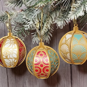 May include: Three spherical Christmas baubles in gold, each with detailed cut-out patterns. The colour schemes are red and white, red and teal, and teal and white. They are suspended by gold ribbons from a frosted evergreen branch.