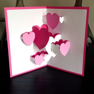 May include: A pink and white pop-up card with a 3D design of multiple hearts. The card is open and the hearts are layered on top of each other.