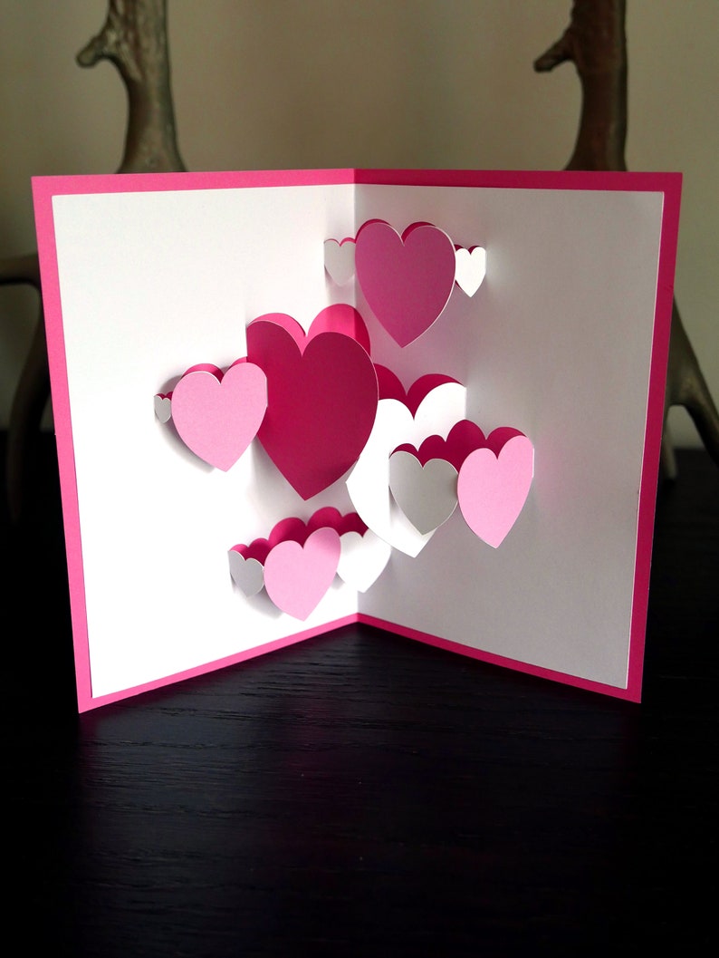 May include: A pink and white pop-up card with a 3D design of multiple hearts. The card is open and the hearts are layered on top of each other.