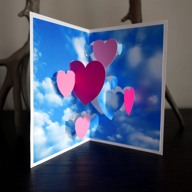May include: A pop-up card with a blue sky and white clouds background. The card features multiple pink and blue paper hearts in a 3D design.