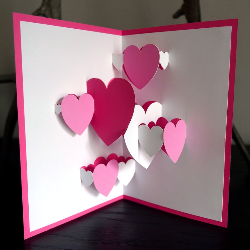 May include: A pink and white pop-up card with a 3D design of multiple hearts. The card is open and the hearts are layered in a cascading effect.