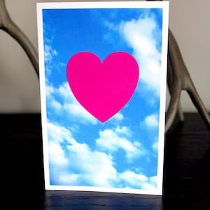 May include: A greeting card with a bright pink heart shape against a blue sky with white clouds.