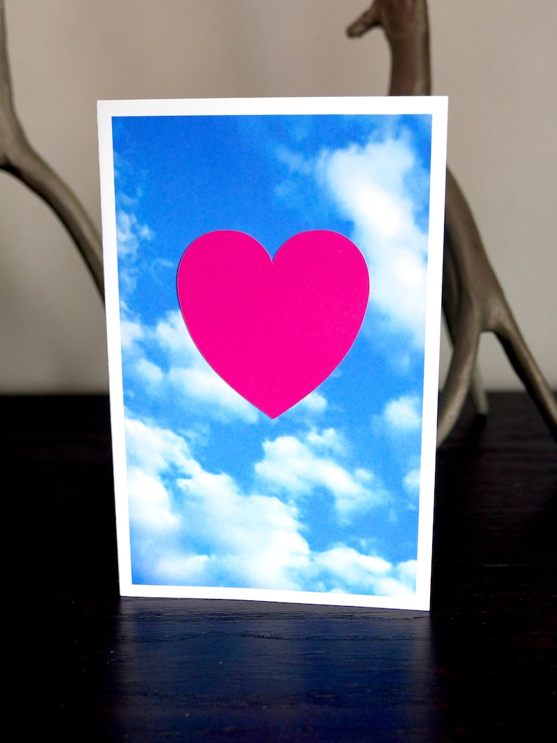 May include: A greeting card with a bright pink heart shape against a blue sky with white clouds.
