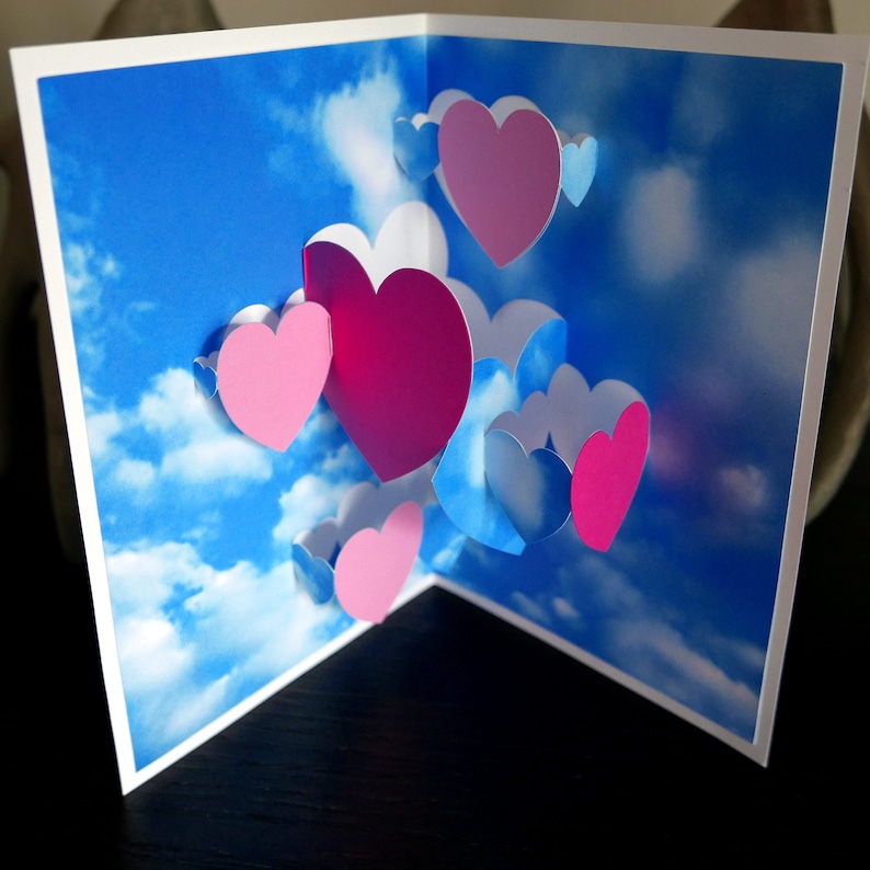 May include: A 3D pop-up card with a blue sky and white clouds background. The card features multiple pink and blue hearts in a layered design.