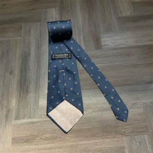 Puede incluir: Una corbata azul oscuro con un patrón repetitivo de pequeños cuadrados blancos y rojos. La corbata incluye una etiqueta negra con el texto Christian Dior Made in U.S.A. y un forro de color claro estampado con el nombre de la marca Christian Dior.