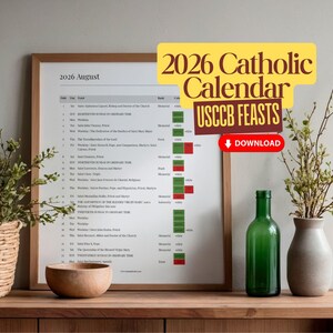 May include: A framed 2026 Catholic calendar with USCCB Feasts, displayed on a wooden surface. The calendar is next to a green glass bottle, a beige vase with greenery, a woven basket with greenery, and a wooden bowl.