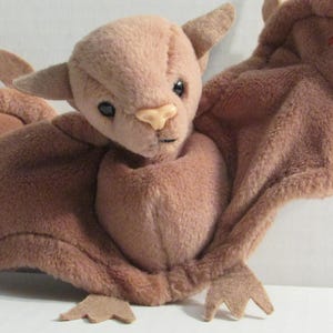 Könnte beinhalten: Ein Plüschtier in Form einer Fledermaus mit weicher, brauner Fellstruktur. Die Fledermaus hat große, ausgebreitete Flügel, kleine Ohren und ein detailliertes Gesicht mit schwarzen Augen und einer hellen Nase. Die Füße der Fledermaus sind sichtbar.