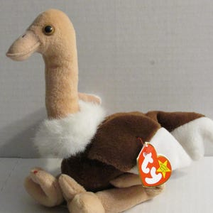 Könnte beinhalten: Plüsch-Strauß mit beigem Körper, braunen Flügeln und weißen Akzenten. Das Spielzeug hat einen langen Hals, einen kleinen Schnabel und ein rotes Etikett. Der Strauß sitzt.