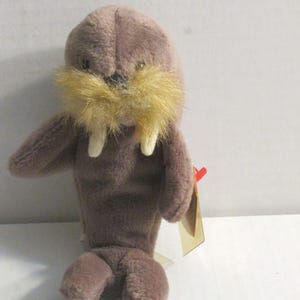 Könnte beinhalten: Ein braunes Plüschtier-Walross mit einem beigefarbenen Schnurrbart und zwei weißen Stoßzähnen. Das Stofftier hat kleine Arme und flossenartige Füße. Ein goldenes Etikett ist am Spielzeug befestigt.