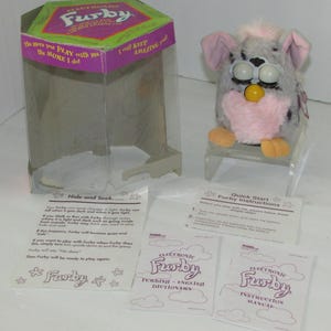 May include: A gray and pink electronic Furby toy with large eyes and a yellow beak, accompanied by its original hexagonal packaging box, a quick start guide, a Furby dictionary, and an instruction manual, all featuring printed text.