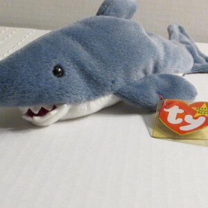 Könnte beinhalten: Ein blaues Plüschhai-Spielzeug mit weißem Bauch und rot ausgekleidetem Maul. Der Hai hat schwarze Augen und Flossen. Ein herzförmiges Etikett in Rot und Gelb ist am Spielzeug befestigt.