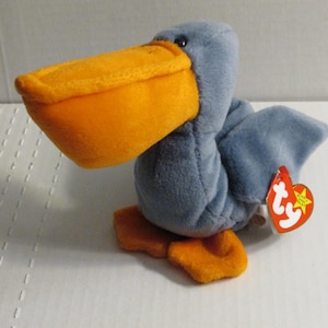 Könnte beinhalten: Ein Plüschpelikan-Spielzeug mit einem großen orangefarbenen Schnabel und Füßen sowie einem blauen Körper und Flügeln. Das Spielzeug hat ein rotes Etikett. Der Pelikan ist ein Stofftier.