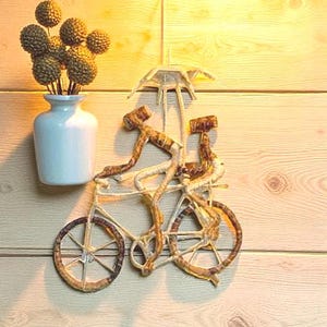 May include: A decorative wall hanging featuring a bicycle with two riders under an umbrella. The bicycle is made of a light brown material with a textured appearance. A light blue vase holds a bouquet of brown, spherical flowers.