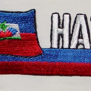 May include: Embroidered patch featuring the flag of Haiti with the word "HAITI" in white, block letters. The flag is red, white, and blue, with a gold pole and accents. The patch is likely for clothing or accessories.