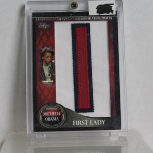 May include: A commemorative card featuring Michelle Obama, labelled "First Lady." The card has a red, white, and blue design with a fabric patch. The card is encased in a clear protective holder.