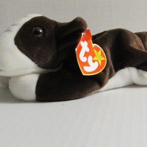 Könnte beinhalten: Ein braun-weißes Stofftier einer Kuh in liegender Position. Das Spielzeug hat ein weißes Gesicht und weiße Beine, mit braunen Flecken auf Körper und Ohren. Ein rotes Etikett mit gelbem Stern ist angebracht.