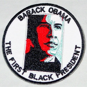 May include: A white circular patch with black embroidered text that reads "Barack Obama The First Black President". The centre of the patch features a black and white portrait of Barack Obama with a red and green overlay.