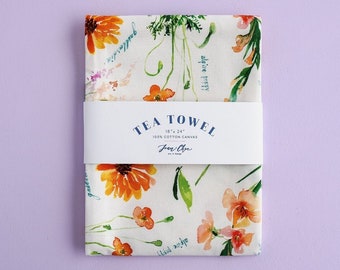 May include: A white tea towel with a floral pattern featuring orange, yellow, and green watercolor flowers. The text "TEA TOWEL 18" x 24" 100% COTTON CANVAS Jean & Co. Art & Design" is printed on a white label attached to the towel.