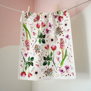 May include: A white tea towel with a floral pattern featuring pink roses, white anemones, and green leaves. The flowers are surrounded by small red berries and the names of the flowers are written in black ink.