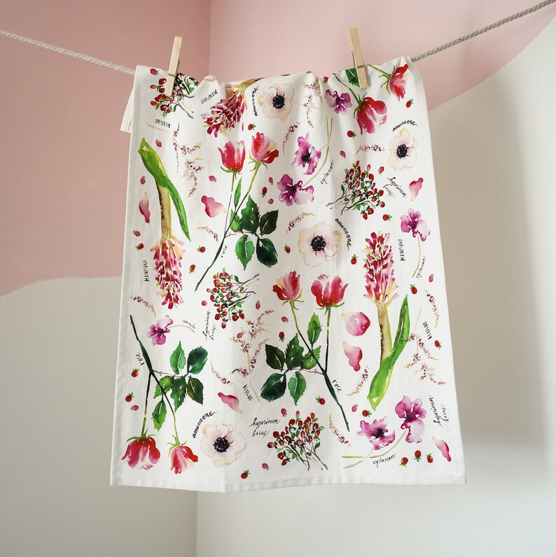 May include: A white tea towel with a floral pattern featuring pink roses, white anemones, and green leaves. The flowers are surrounded by small red berries and the names of the flowers are written in black ink.