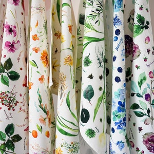May include: Set of seven white tea towels with watercolor floral designs. Each towel features a different floral pattern, including berries, flowers, and leaves. The towels are hung on a white wall.