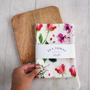 May include: A white tea towel with a floral pattern featuring pink and red flowers, green leaves, and small red berries. The towel has a label that reads "TEA TOWEL 18" x 24" 100% Cotton Canvas Jane Chen".