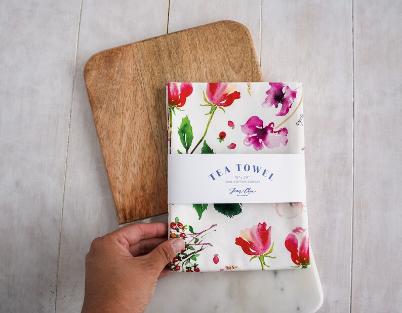 May include: A white tea towel with a floral pattern featuring pink and red flowers, green leaves, and small red berries. The towel has a label that reads "TEA TOWEL 18" x 24" 100% Cotton Canvas Jane Chen".