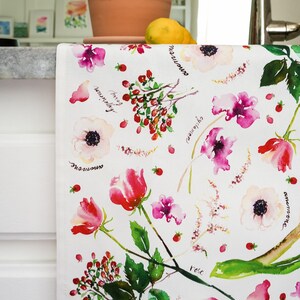 May include: White kitchen towel with a watercolor floral pattern. The flowers are pink, red, and white. The names of the flowers are written in black ink on the towel. The flowers include rose, anemone, and sweet pea.