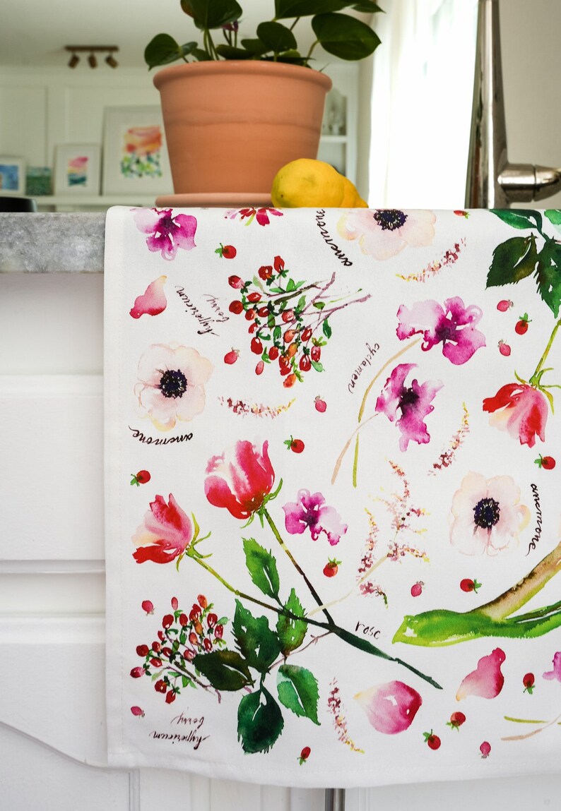 May include: White kitchen towel with a watercolor floral pattern. The flowers are pink, red, and white. The names of the flowers are written in black ink on the towel. The flowers include rose, anemone, and sweet pea.