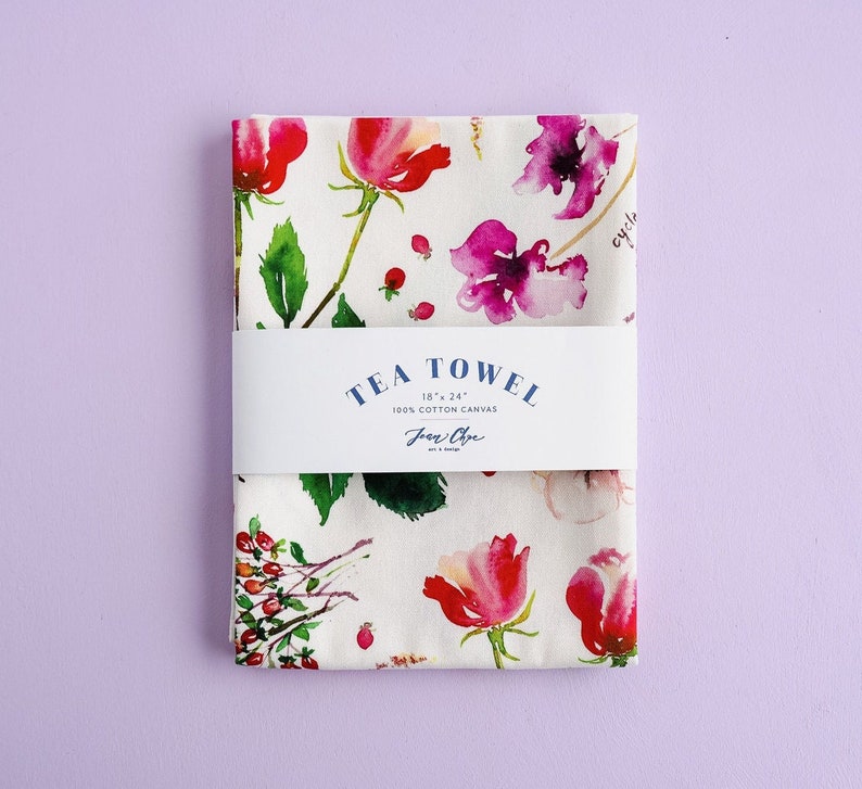 May include: White tea towel with a floral pattern of pink and purple flowers and green leaves. The towel has the text "TEA TOWEL 18" x 24" 100% COTTON CANVAS Jean Cline art & design" printed on it.