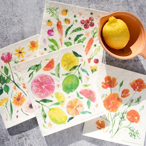 May include: Four square cellulose sponges with different watercolor designs. The designs include a floral pattern, a citrus pattern, a vegetable pattern, and a poppy pattern. The sponges are on a gray surface with a small bowl and a lemon.