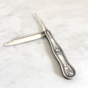 May include: A silver-colored, antique pocket knife with an ornate floral design on the handle. The knife blade and pick are extended, showcasing the details of the vintage design. The knife is set against a white marble surface.