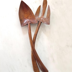 May include: A pair of wooden salad servers, including a spoon and a fork. The spoon has a spade-like head, and the fork has three tines. Both are made of a dark brown wood and are intertwined, creating a decorative and functional kitchen accessory.