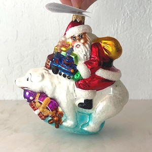 May include: A Christmas ornament featuring Santa Claus riding a polar bear. Santa is dressed in a red suit with a white beard and a gold sack. The polar bear is white with a blue base, and the ornament includes wrapped gifts and a toy train.