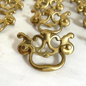 May include: A collection of gold-colored metal drawer pulls featuring a decorative backplate with an ornate, symmetrical cutout design and a curved, swinging handle. Each piece has a polished finish and a distinct, intricate shape for furniture.