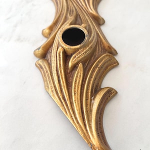 May include: Ornate, gold-colored metal decorative piece with a central circular opening. The design features a stylized, flowing pattern of leaves and curves, with a slightly aged appearance. The piece is likely a decorative element.