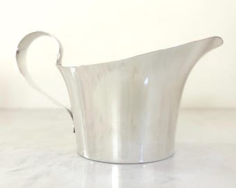 May include: A polished silver-toned gravy boat featuring a smooth, curved body with a wide pouring spout and a single, slender handle extending from the side. The metallic surface has a reflective finish and a simple, clean silhouette.