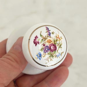 May include: A round white ceramic cabinet knob featuring a central floral pattern with purple, yellow, and blue flowers surrounded by green leaves. The circular face of the knob is bordered by a thin gold metallic rim, providing a decorative accent.