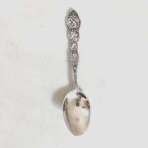 May include: A silver spoon with an ornate handle featuring detailed relief carvings. The spoon's bowl is oval-shaped and polished, reflecting the surrounding environment. The handle's design includes figures and decorative patterns, suggesting a vintage or antique style.