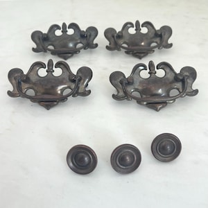 May include: A set of four dark metal drawer pulls with ornate, symmetrical backplates and swinging bail handles, accompanied by three matching circular metal knobs. Each piece features a smooth, darkened finish with subtle highlights on the edges.