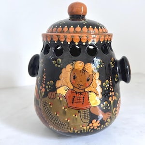 May include: A black ceramic jar with a lid, featuring a hand-painted design of a girl in traditional clothing. The jar has a series of circular cutouts and two side handles. The lid is topped with a brown knob.