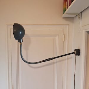 May include: A grey adjustable wall lamp with a round shade and a long, flexible arm. The lamp is mounted on a white wall and has a cord hanging down.