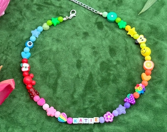 May include: A rainbow beaded necklace with a silver clasp and a name charm that says "KATIE". The necklace features various colorful beads, including hearts, stars, smiley faces, and fruit shapes.