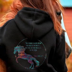 May include: Black equestrian hoodie with a colorful horse graphic and text. The hoodie features a drawstring hood and a kangaroo pocket. The text on the back reads "The horse is made ready for the day of battle, but the victory belongs to the Lord."