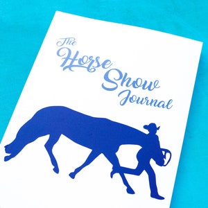 May include: A white journal with a blue silhouette of a horse and rider. The text "The Horse Show Journal" is printed on the cover.