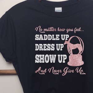 May include: Black t-shirt with a pink glitter design that says "No matter how you feel... Saddle Up Dress Up Show Up And Never Give Up."