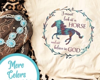 May include: A beige t-shirt with a graphic of a colorful horse and a wreath with the text "I cannot look at a HORSE and not believe in GOD". The shirt is on a wooden surface with a pair of brown cowboy boots, a leopard print scarf, and blue jeans.