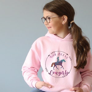 May include: A pink hoodie with a graphic of a girl riding a horse in a circle with the text "God Bless Ya'll Cowgirls".