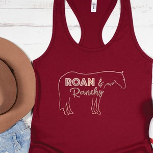 May include: A maroon racerback tank top with the words "ROAN & Ranchy" in white, along with a horse outline. A brown hat, denim shorts, and white sneakers with a leopard print interior are also visible.